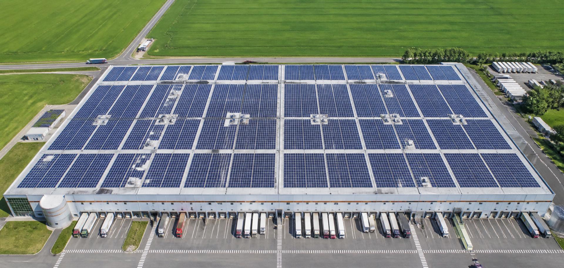 Top down aerial view of a warehouse roof covered in solar panels with loading docks and farmland beyond
